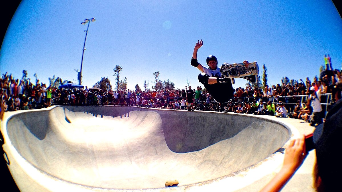 Tony Hawk en plein air au-dessus d’un bowl en béton devant la foule, planche saisie à la main