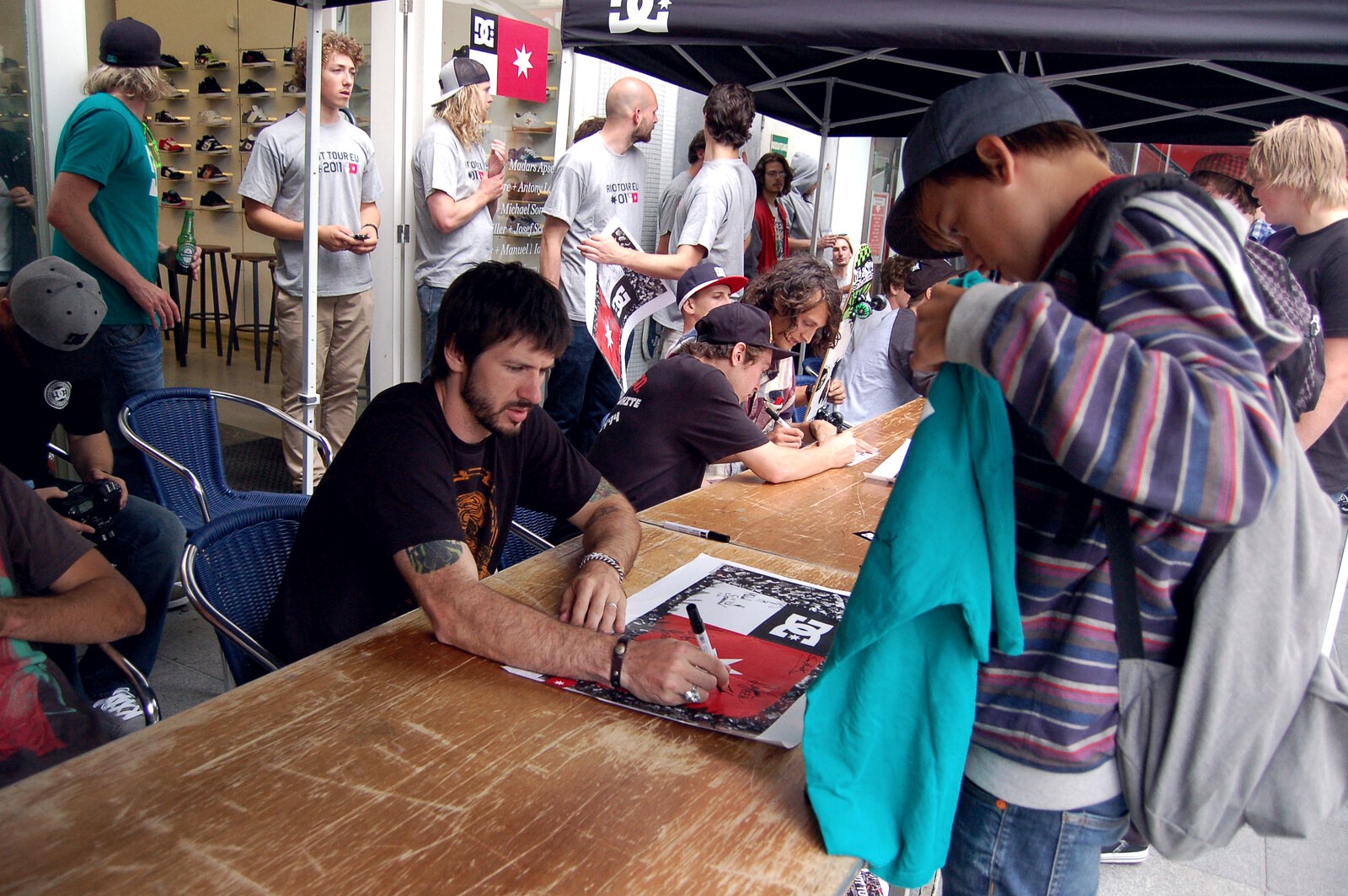 Chris Cole signe des autographes devant un skateshop entouré de skateurs, mur de chaussures visible en arrière-plan