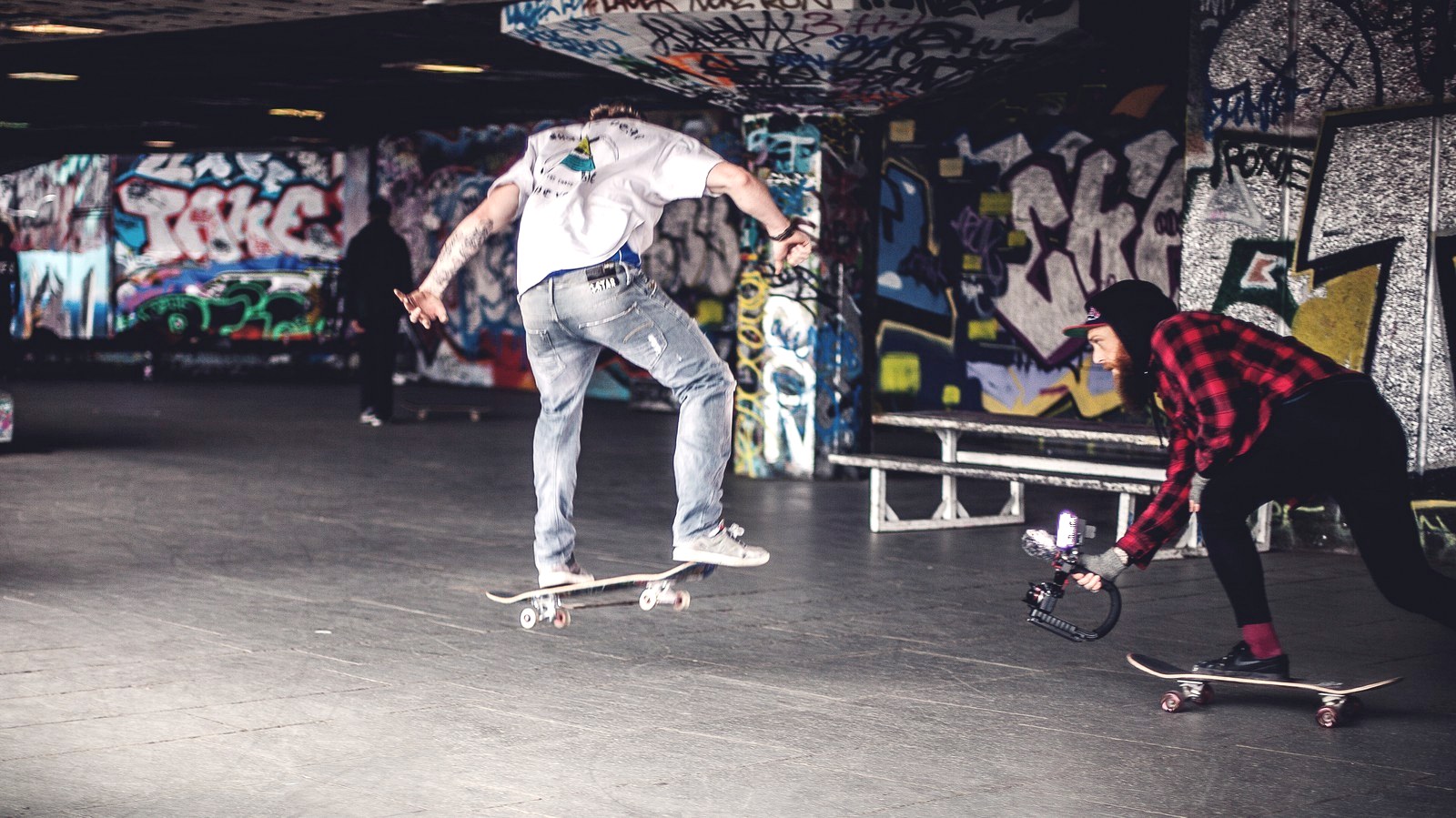 Deux skateurs sur un sol plat en béton sous un pont couvert de graffitis