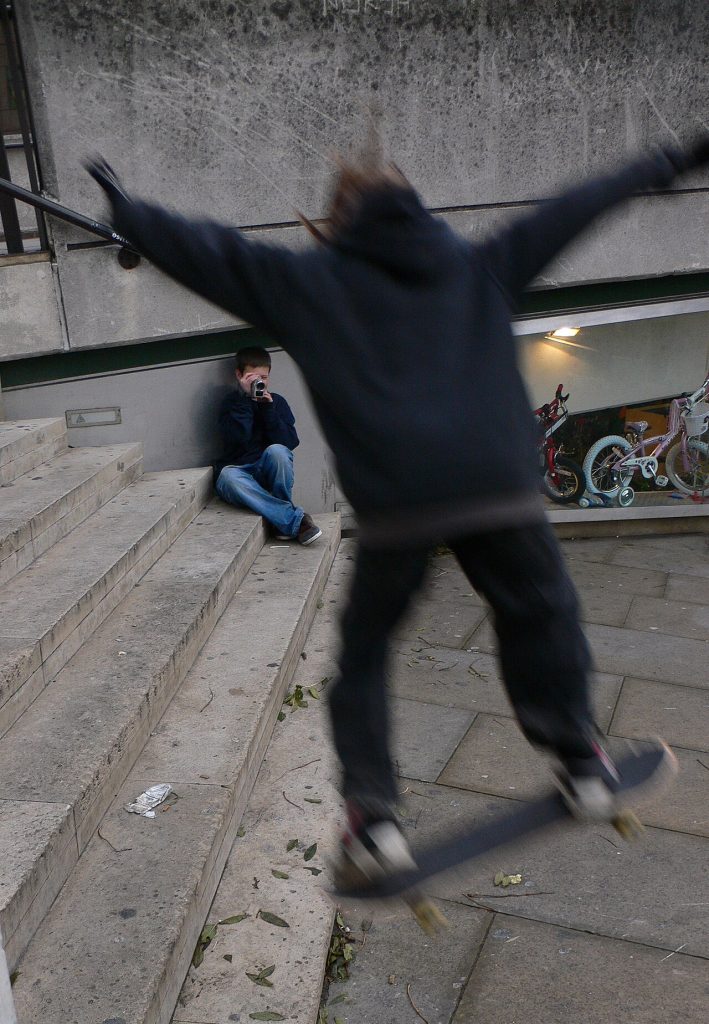 Un gamin assis dans les marches filme au camescope un skateur en plein saut, la camera qui enregistre surtout les rates