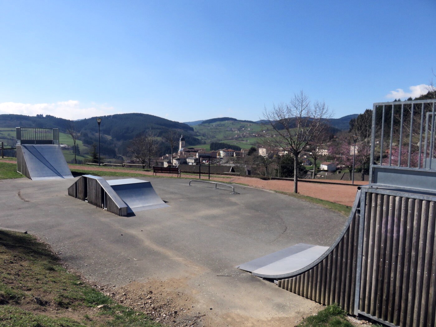 Skatepark de village à Grandris : modules métalliques boulonnés sur une dalle de bitume, clocher et collines au fond, aucun skateur