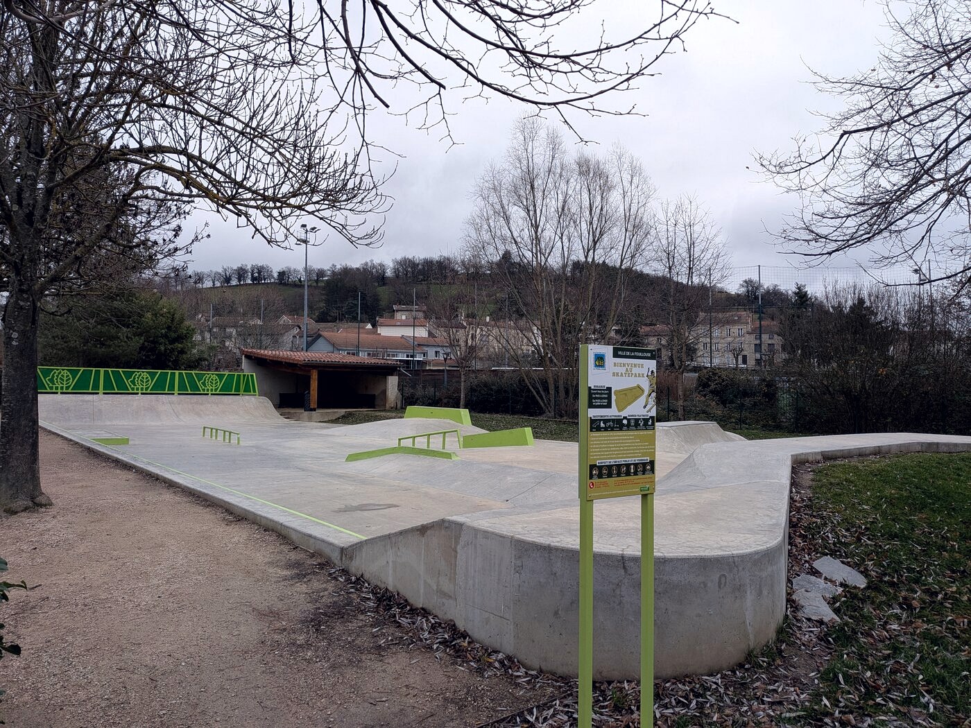 Skatepark en béton récent et désert à La Fouillouse, avec panneau réglementaire et modules verts, un après-midi de décembre