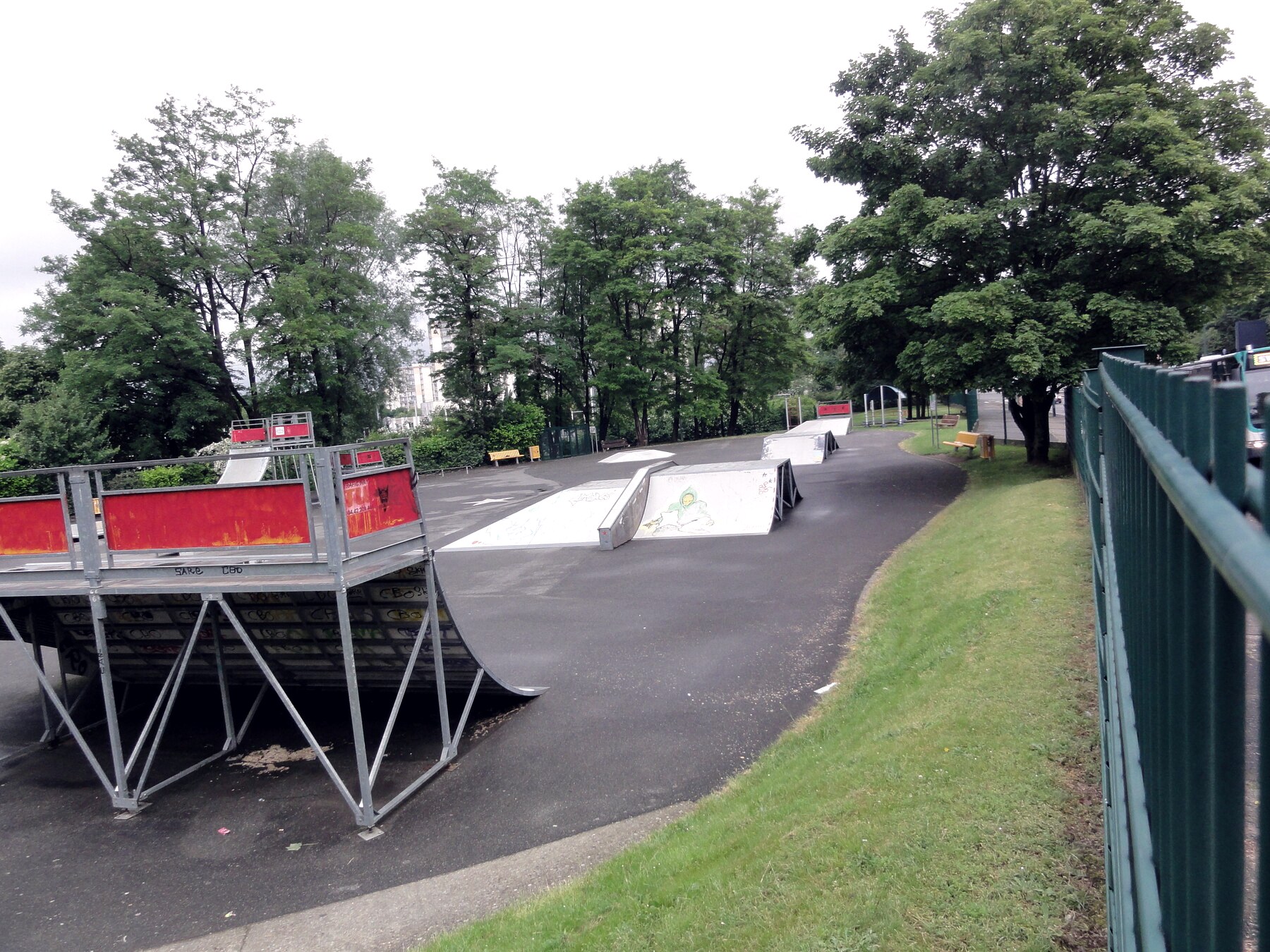 Skatepark municipal français : modules métalliques sur bitume, derrière un grillage vert, à l'écart de la ville