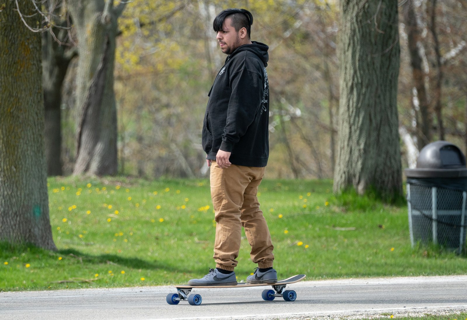 Homme en sweat noir debout sur un cruiser aux roues bleues, pieds posés, qui garde son élan sur l’allée d’un parc