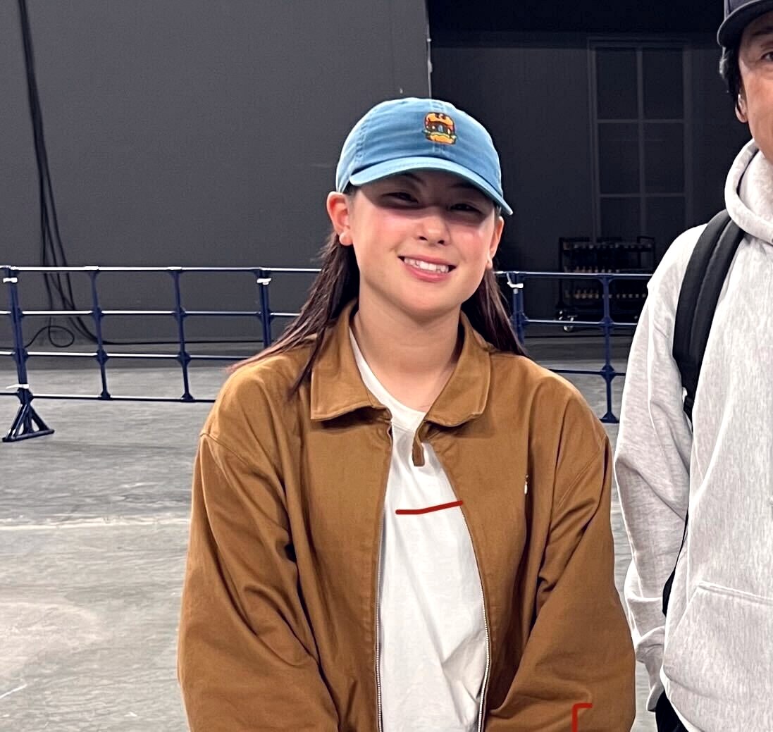 Miyu Ito souriante en veste de travail beige, casquette bleue, dans les coulisses d'une compétition de skate